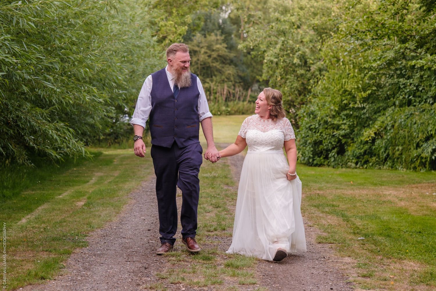 A couple in wedding attire walking together on a rustic outdoor path surrounded by lush green foliage.