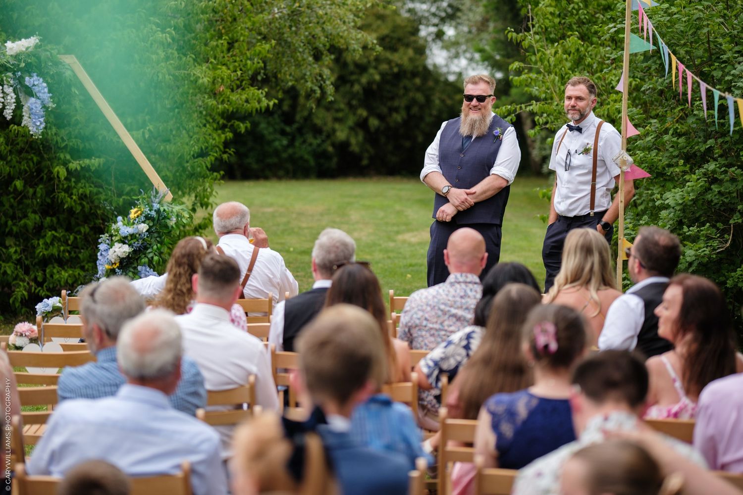 An outdoor garden wedding ceremony with guests sitting on wooden chairs in rows beneath trees and bunting.