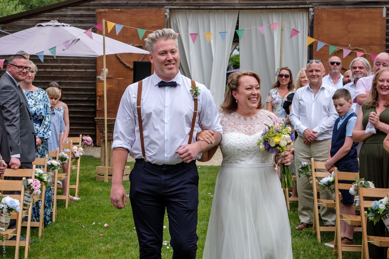 Couple walks down outdoor aisle at backyard garden wedding with guests seated on wooden chairs and umbrellas overhead.