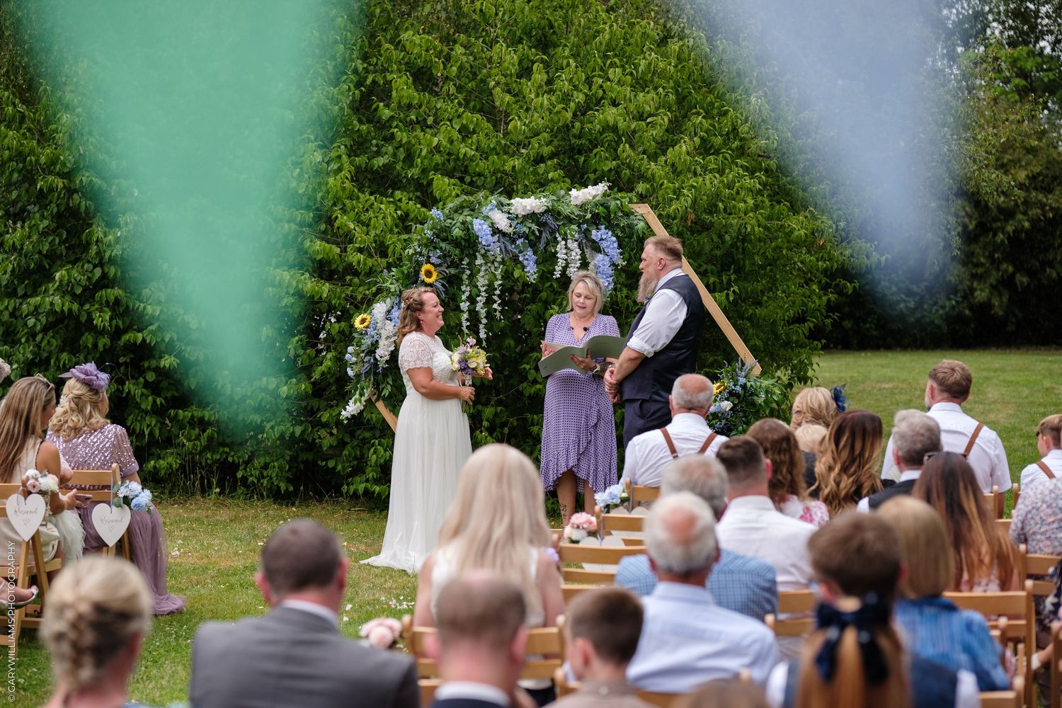 Outdoor garden wedding ceremony with guests seated on wooden chairs beneath a floral-decorated arch on a sunny day.