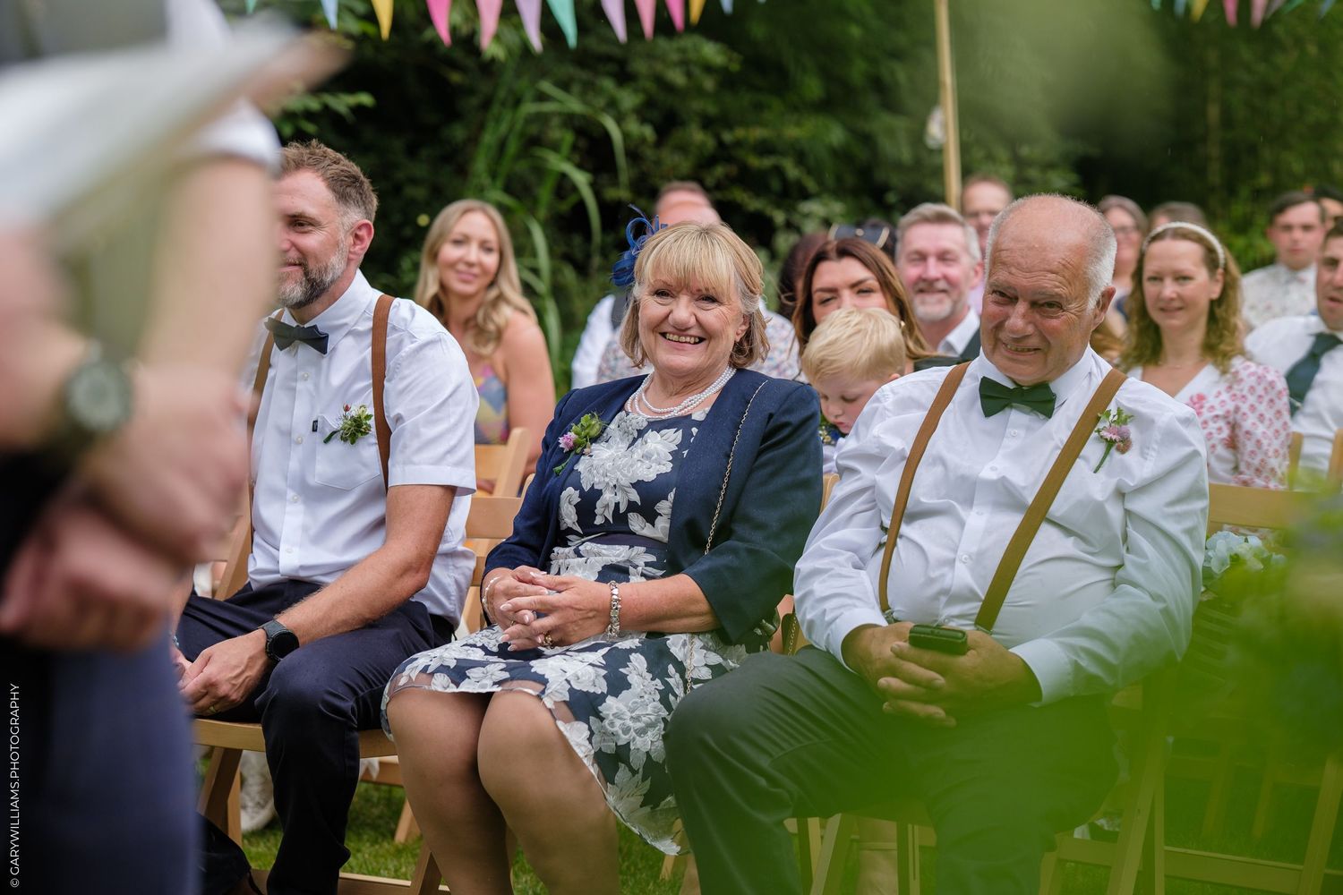Guests are seated outdoors during a garden wedding ceremony with decorative plants in the foreground.