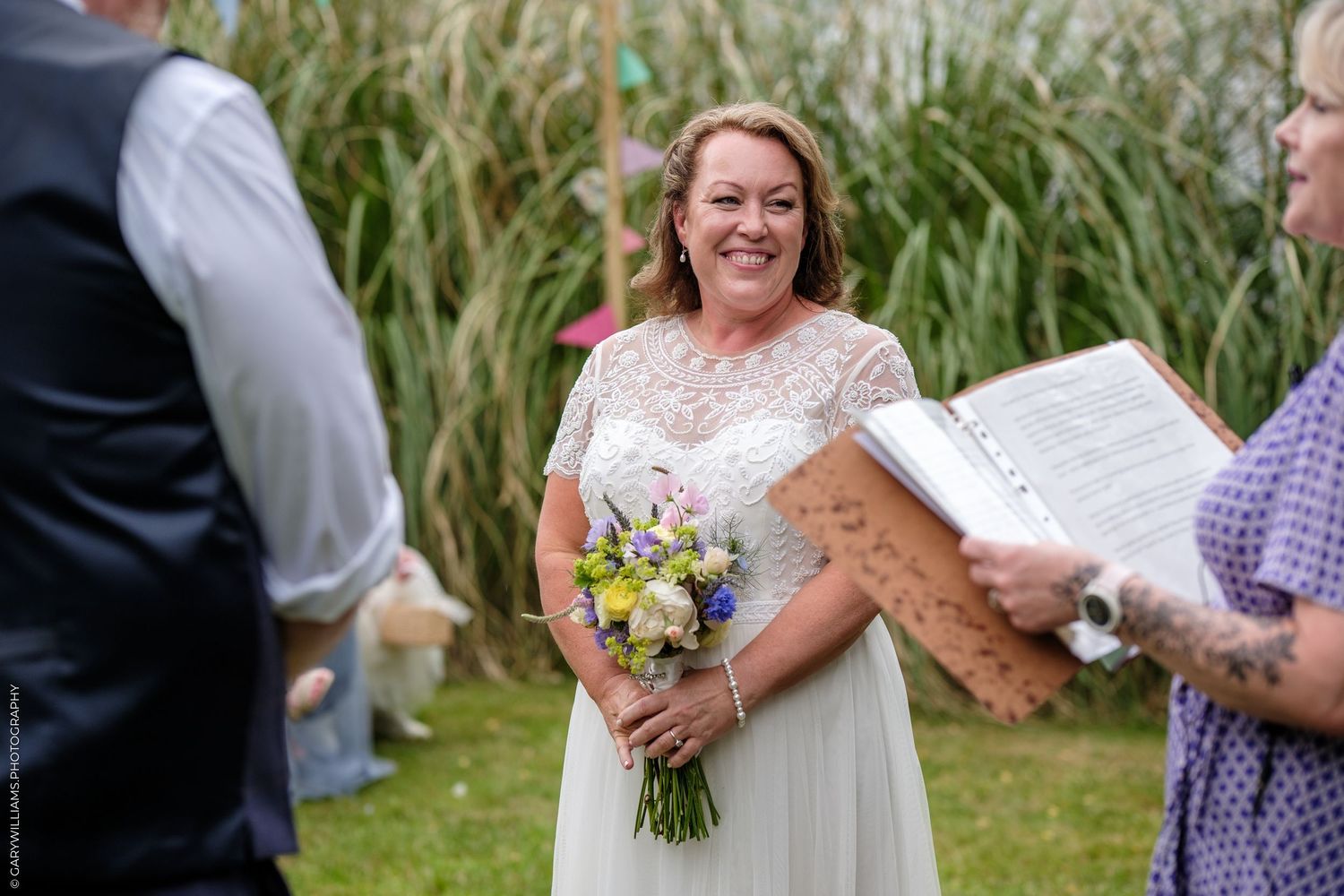 White-clad bride holds yellow and white wildflower bouquet during outdoor garden wedding ceremony.