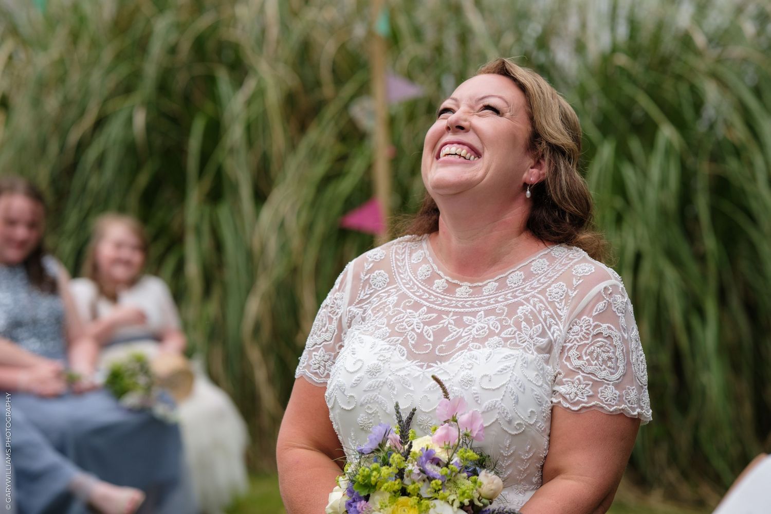 A bride in a white lace gown laughs joyfully while holding a bouquet with greenery in the background.