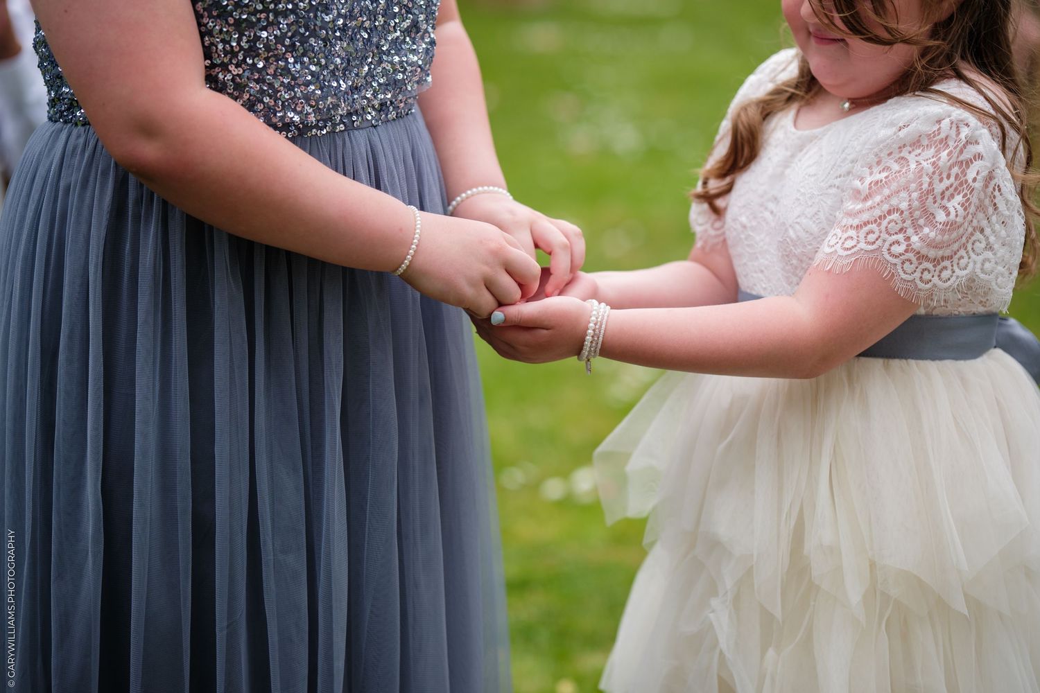 A person in a gray dress holds hands with a small child wearing a white lacy dress with a sash outdoors.