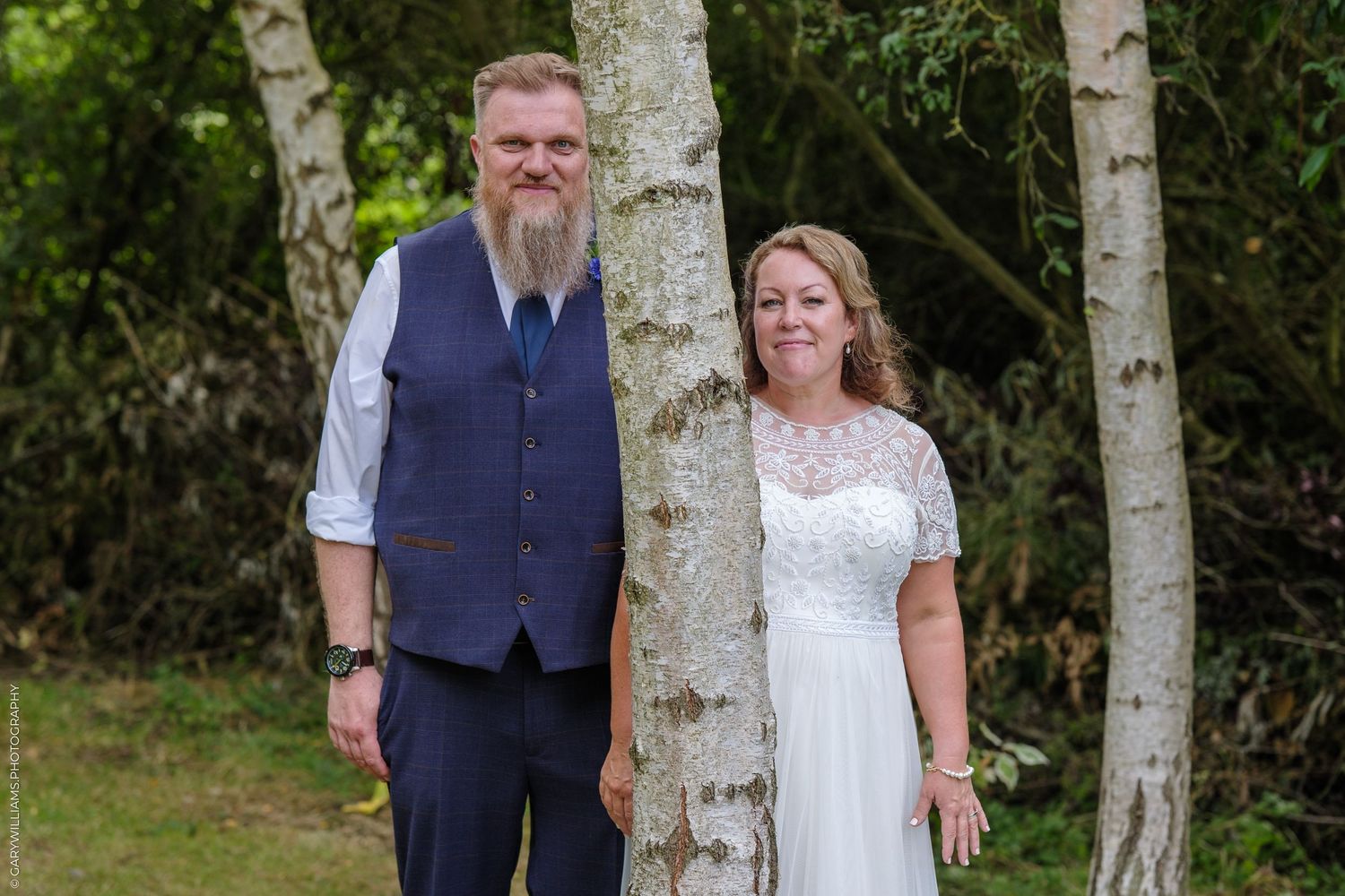 A couple stands together among birch trees at an outdoor wedding, wearing a navy vest and white lace dress.