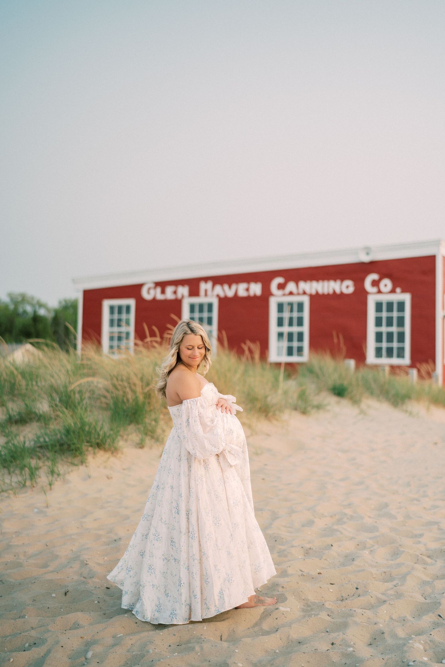 White lace dress flows gracefully on sandy beach with red barn and white windows in background.
