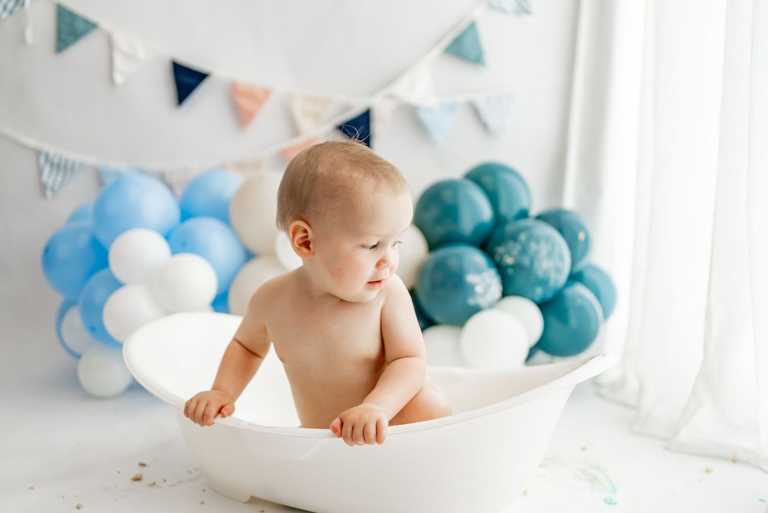 Baby in white bathtub with blue and white balloon decorations during milestone photo session.