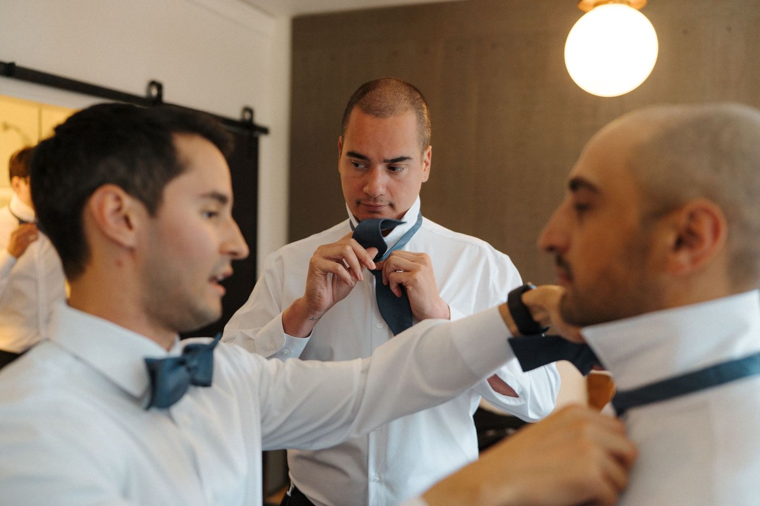 Three people in white shirts adjust and help each other with bow ties while getting ready for a formal event.