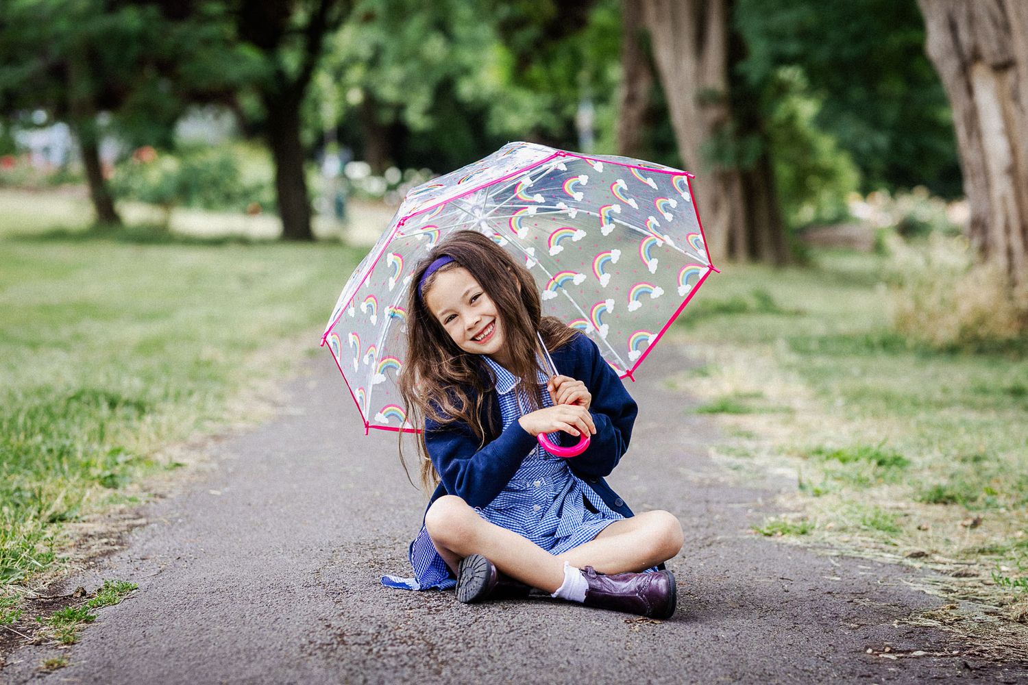A school child sits on a path surrounded by grass and trees. She smiles to camera and holds an umbrella. Bristol school photographer
