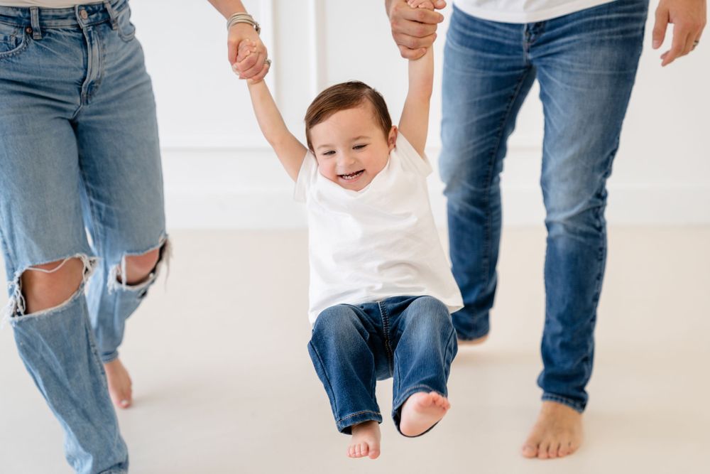 Family wearing denim and white stands barefoot on white studio floor during casual photoshoot.