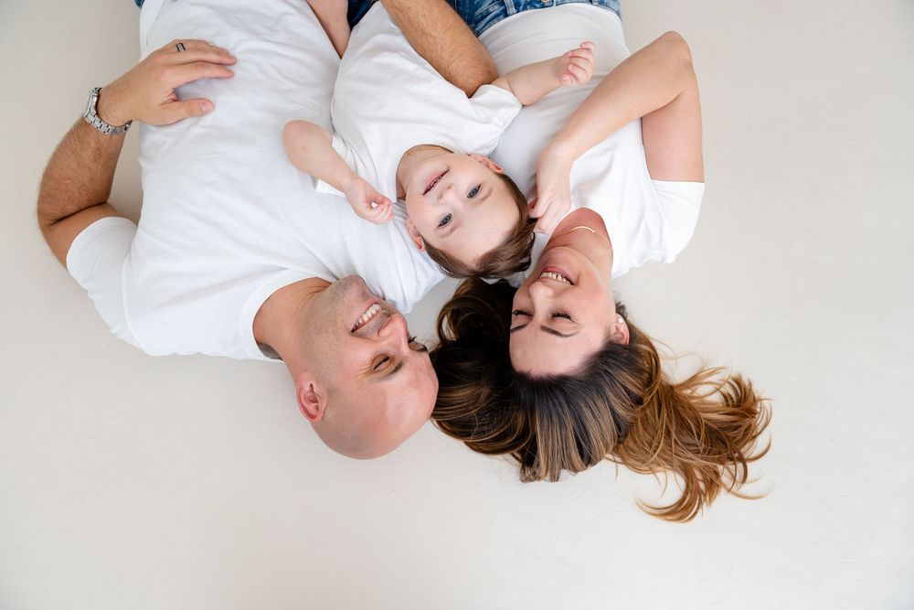 A family lies together on white bedding, forming a circle while smiling and laughing.