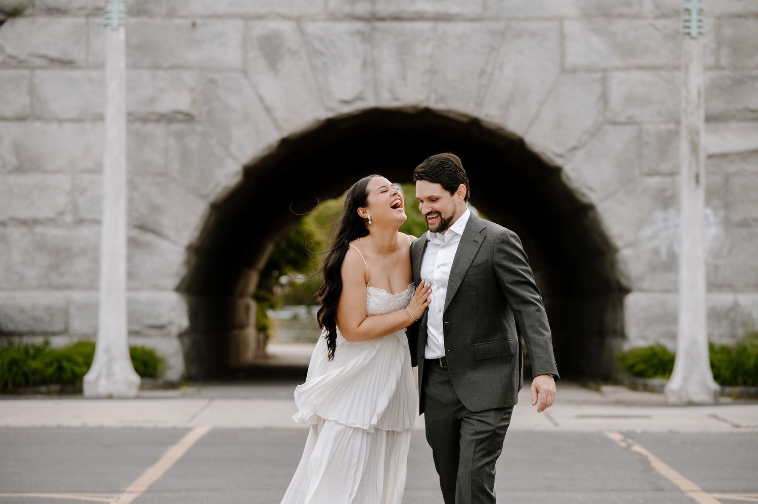 A couple in wedding attire walks together beneath a stone archway while sharing a joyful moment.