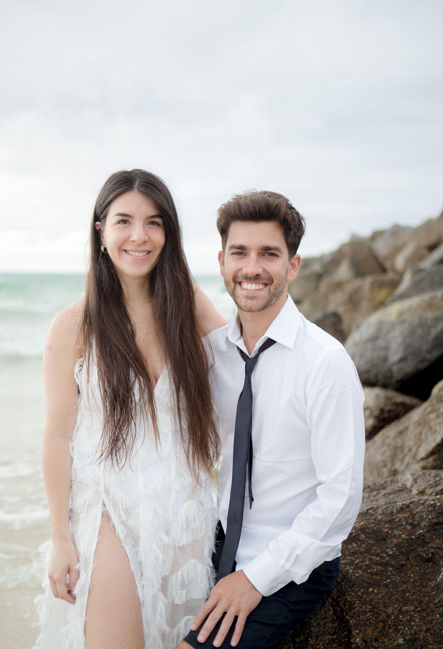 A romantic couple embraces on a sandy beach with stormy waves and rocky coastline in the background.