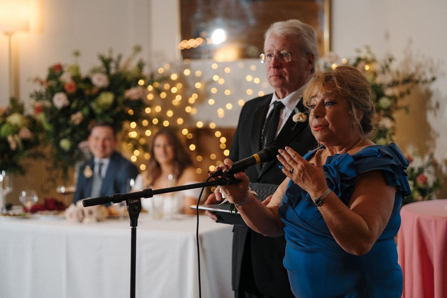 Couple sits at elegant wedding reception table with floral arrangements and twinkling fairy lights in background.