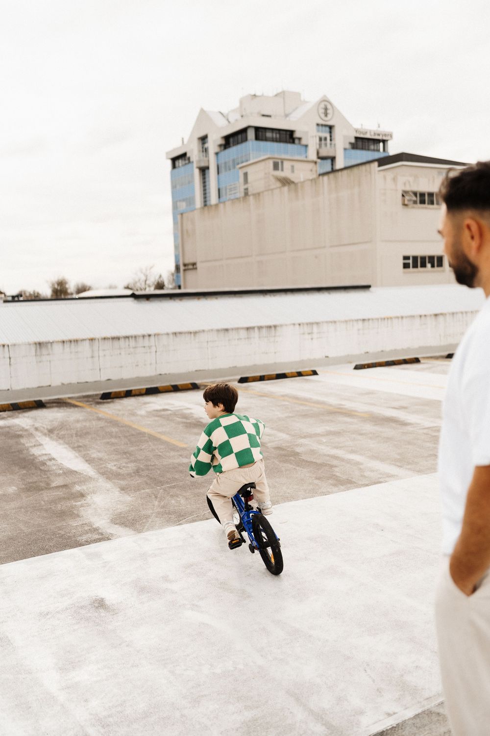 Young child riding a blue bicycle on an empty rooftop parking area while being supervised by an adult.