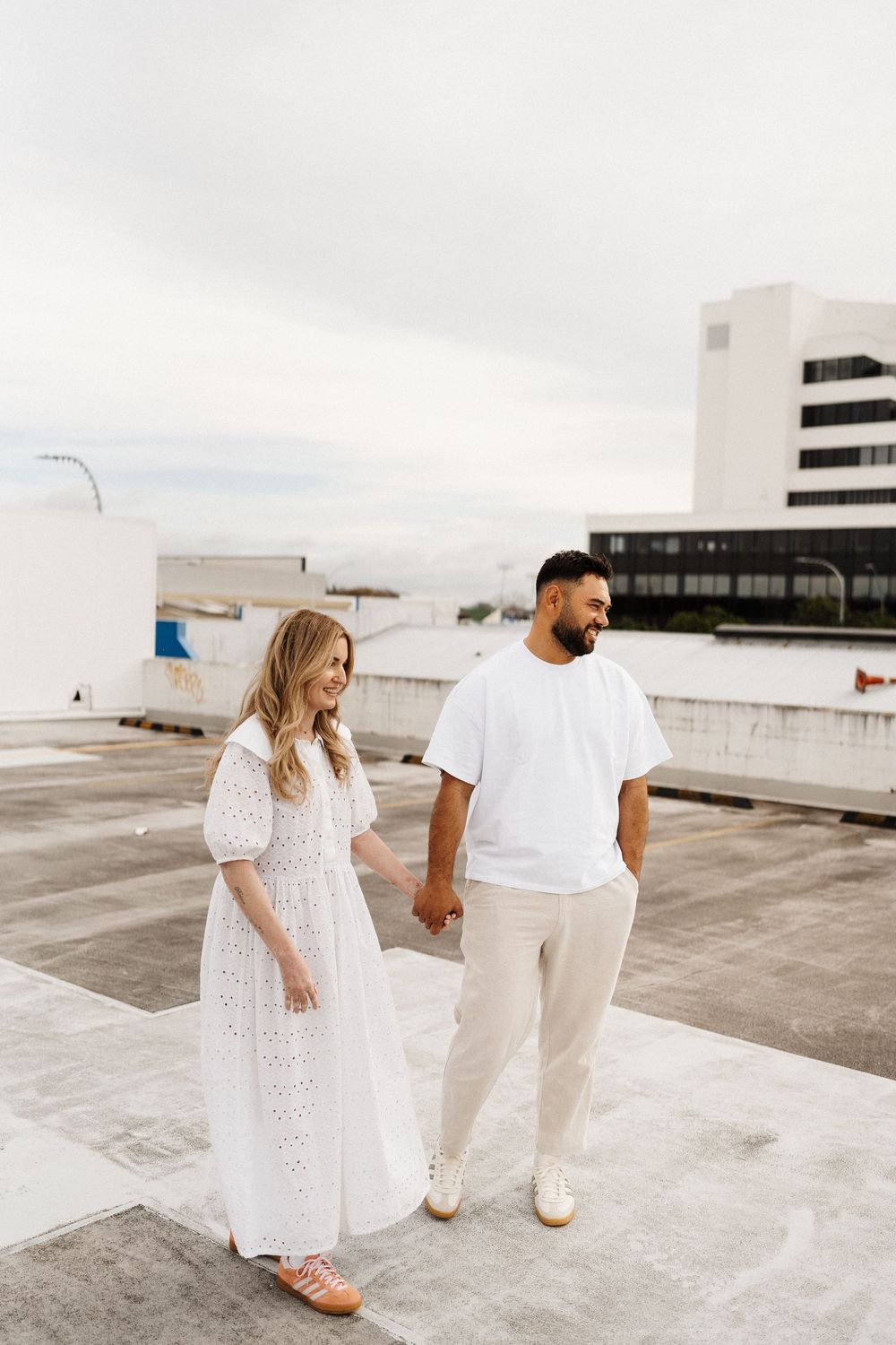 Two people in white outfits hold hands while standing on a rooftop parking structure on an overcast day.