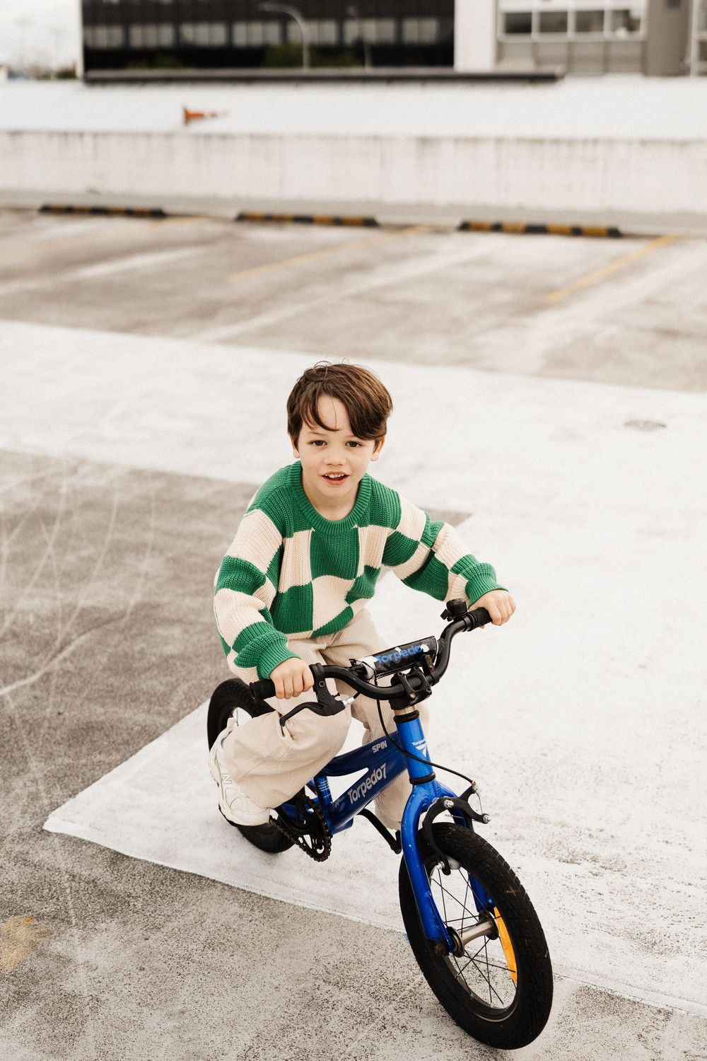 A young child rides a blue and yellow balance bike on a concrete surface while wearing a green and white striped shirt.