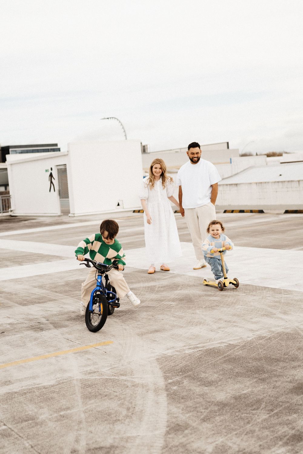 Two children having fun on a rooftop parking lot while their parents watch, one on a bike and one on a skateboard.