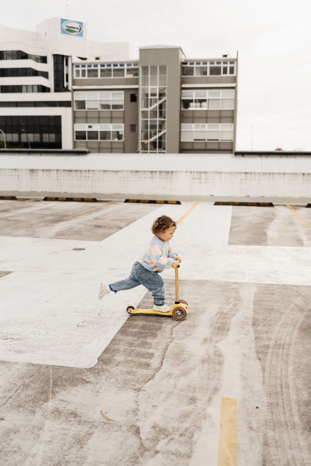 Young child rides yellow kick scooter on concrete rooftop parking area with modern building in background.