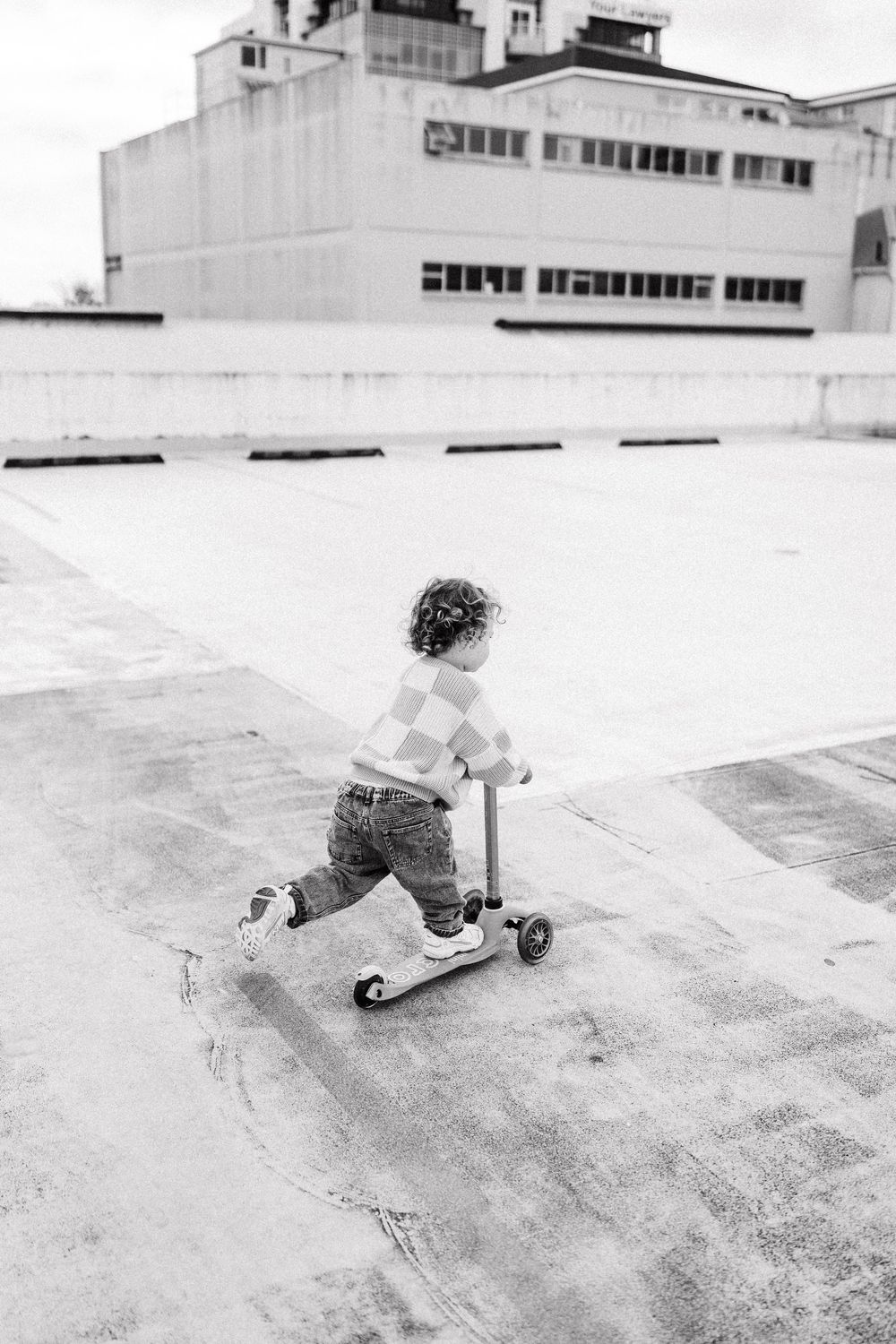 A small child rides a scooter on a rooftop terrace with an industrial building in the background.