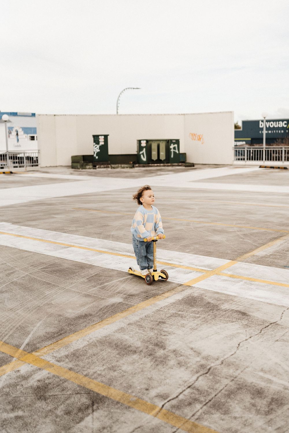 A small child rides a scooter in an empty parking lot with a beige commercial building in the background.