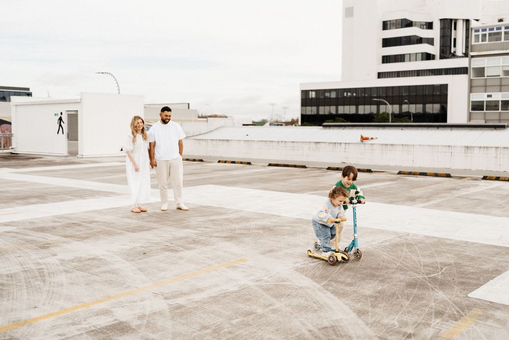 Two people in white outfits watch as a young child rides a tricycle across an empty rooftop parking deck.