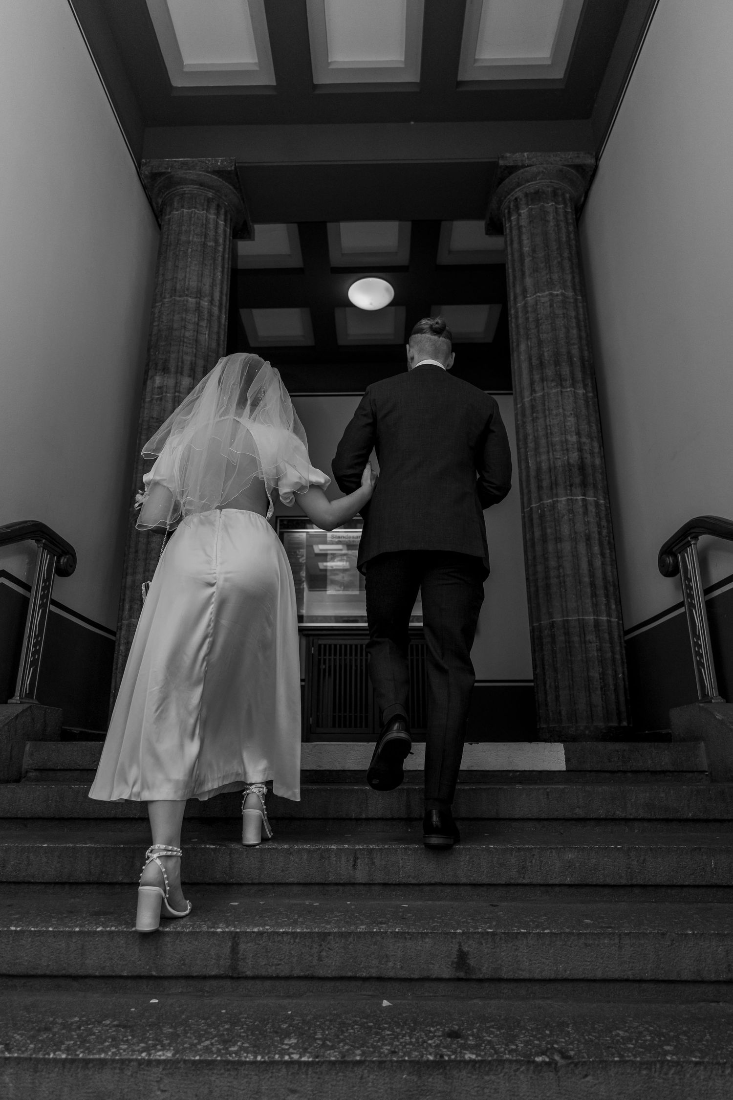 Hochzeitsreportage Standesamt Spandau Berlin – Brautpaar geht Hand in Hand Treppe hinauf Schwarz-Weiß
