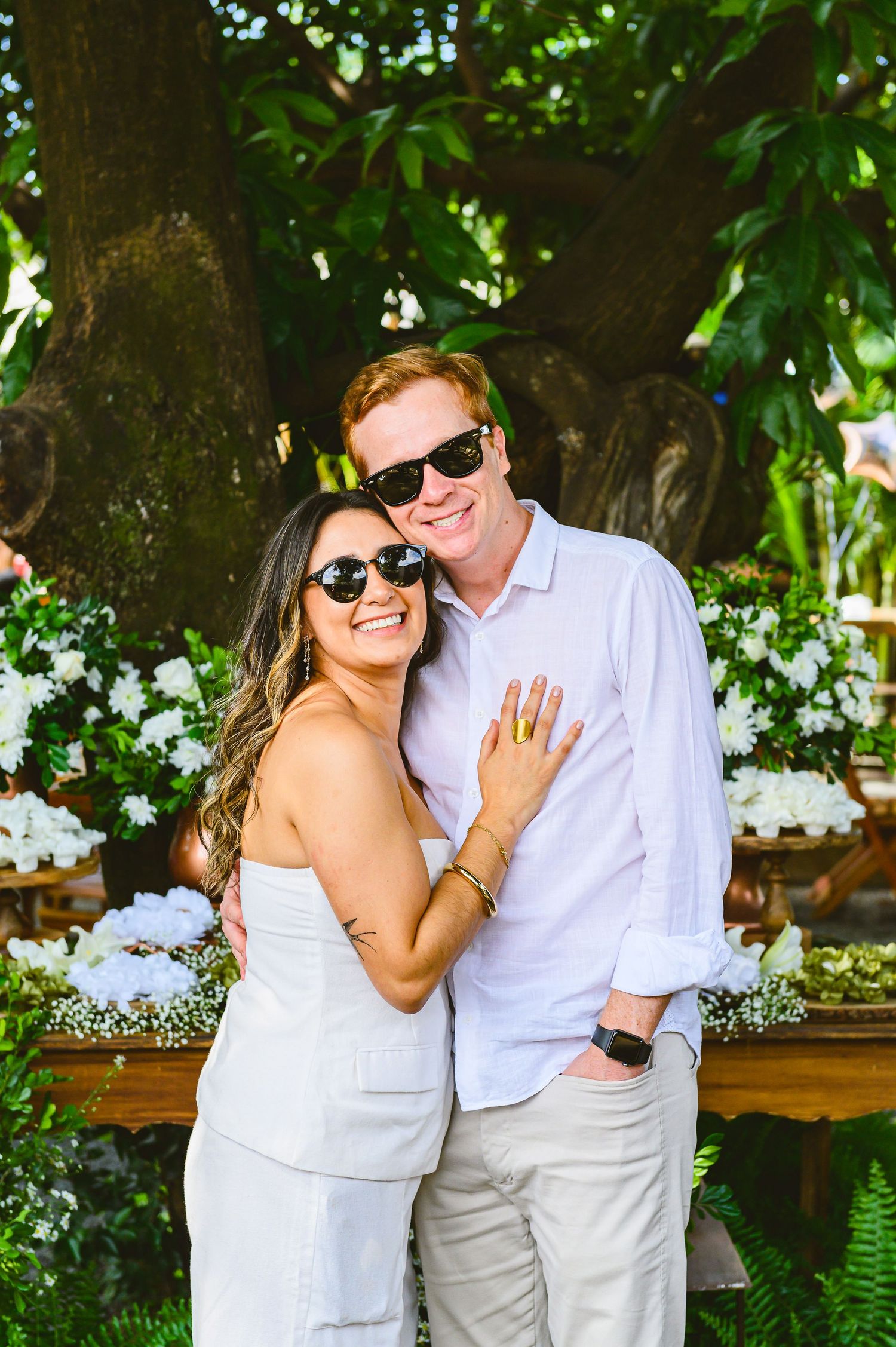 Couple in white attire share joyful moments together in a lush garden setting with flowering plants in the background.