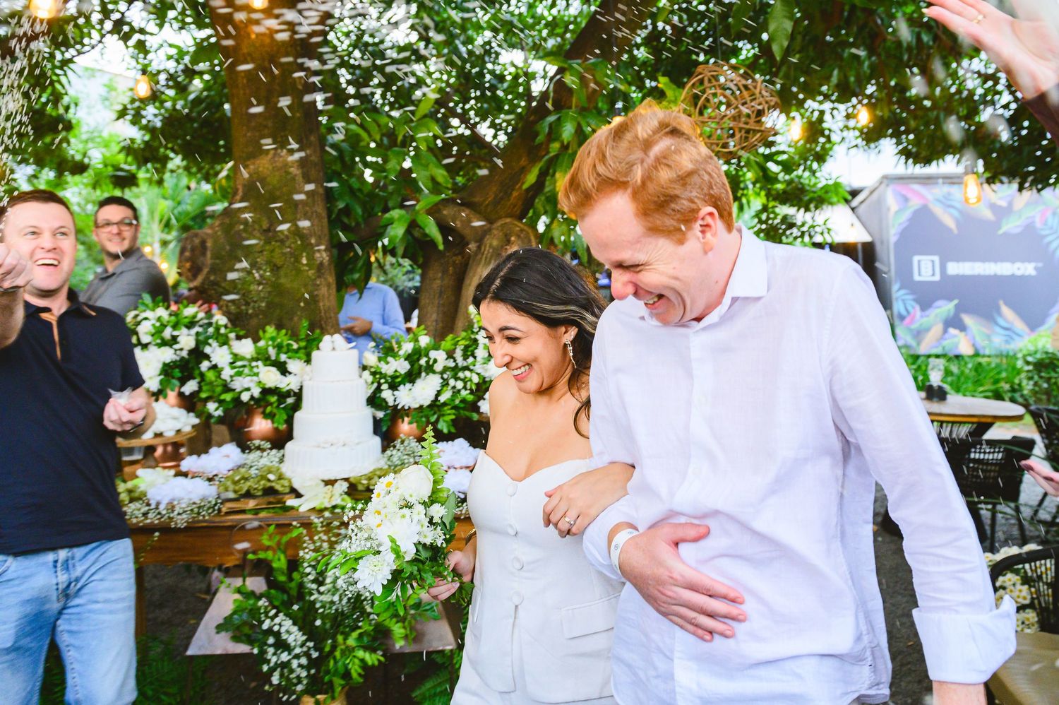 A couple shares a joyful moment under flowering trees at an outdoor celebration with guests nearby.