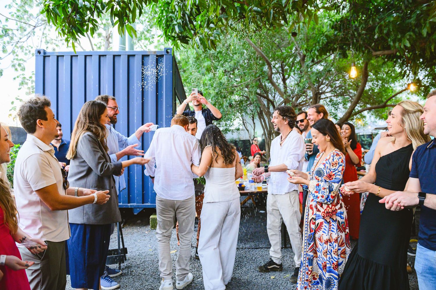 A group of people gathered outdoors for drinks and conversation near a blue shipping container and leafy trees.