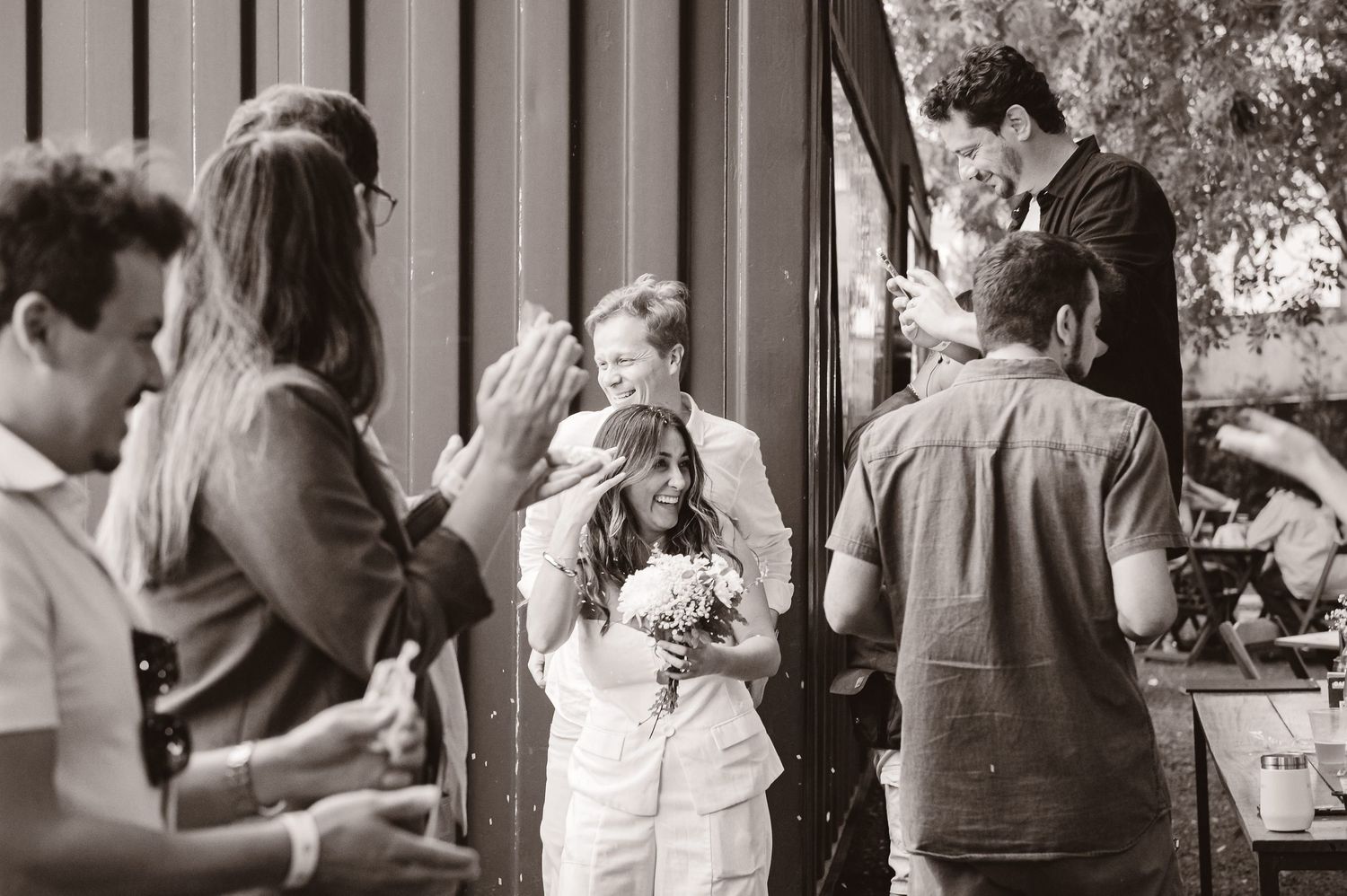 A black and white photograph shows a casual outdoor gathering of people standing and socializing in a vintage setting.