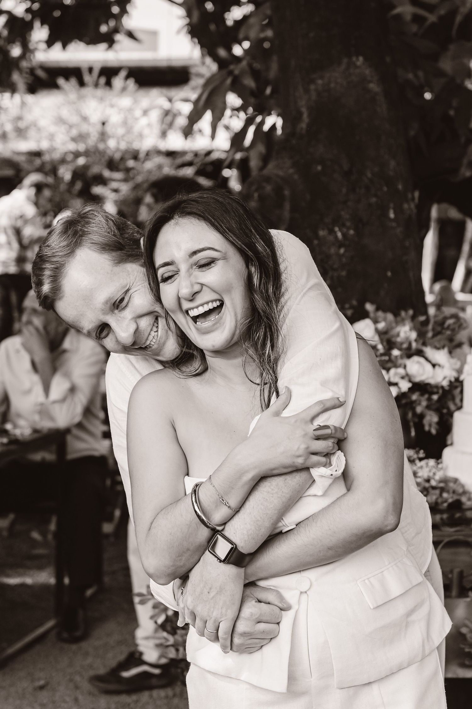 A joyful black and white candid photo capturing an intimate moment of laughter and embracing at an outdoor celebration.