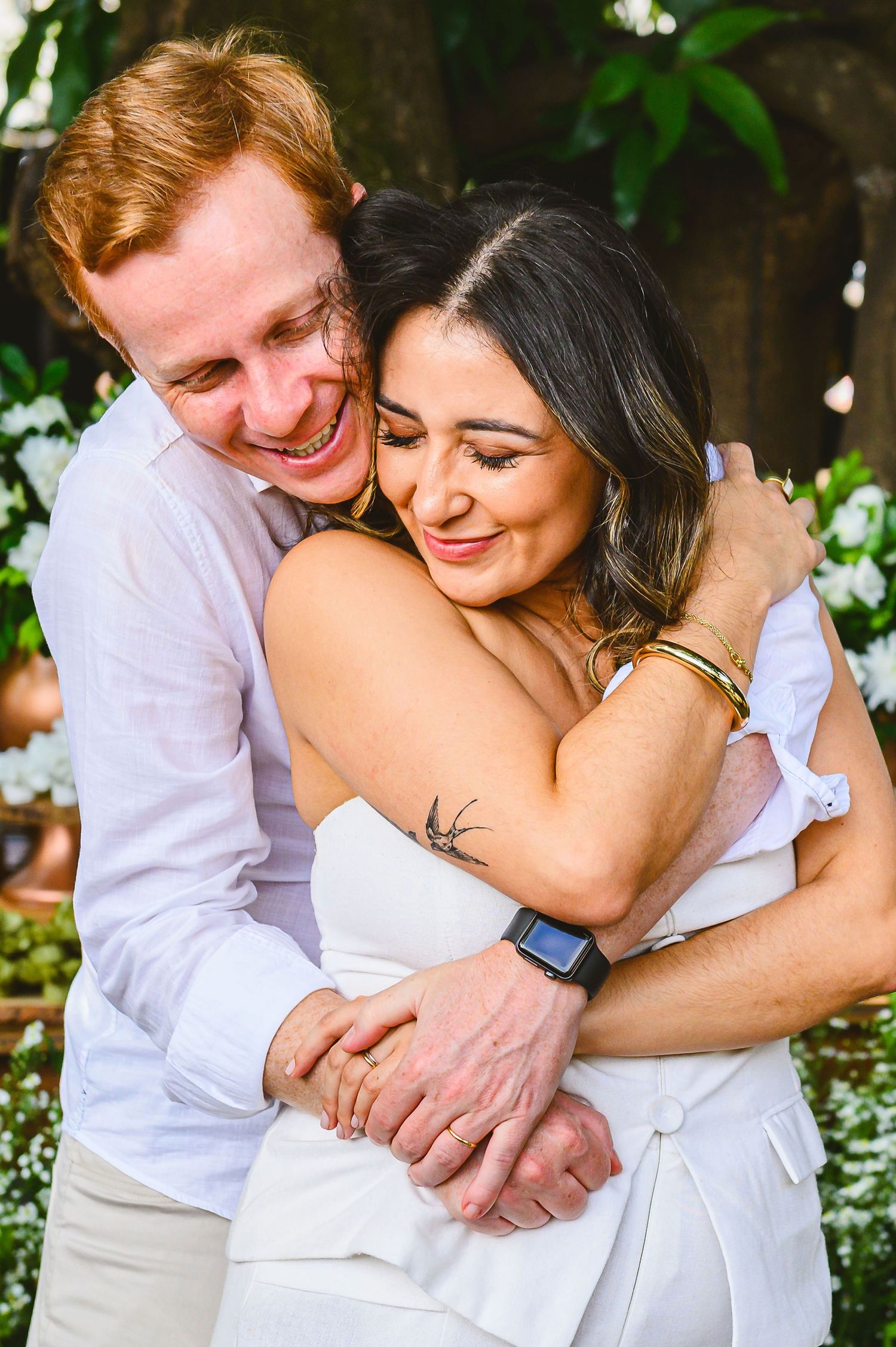 A loving couple embraces and laughs together outdoors among flowering plants on a sunny day.