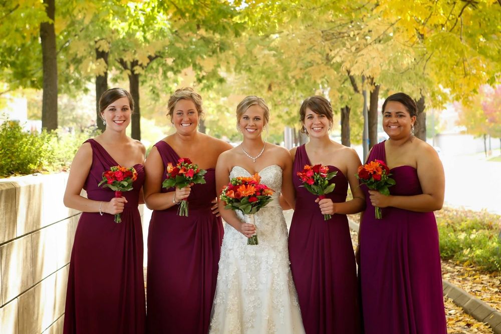 A bridal party in burgundy bridesmaid dresses and white wedding gown pose outdoors on an autumn day with bouquets.