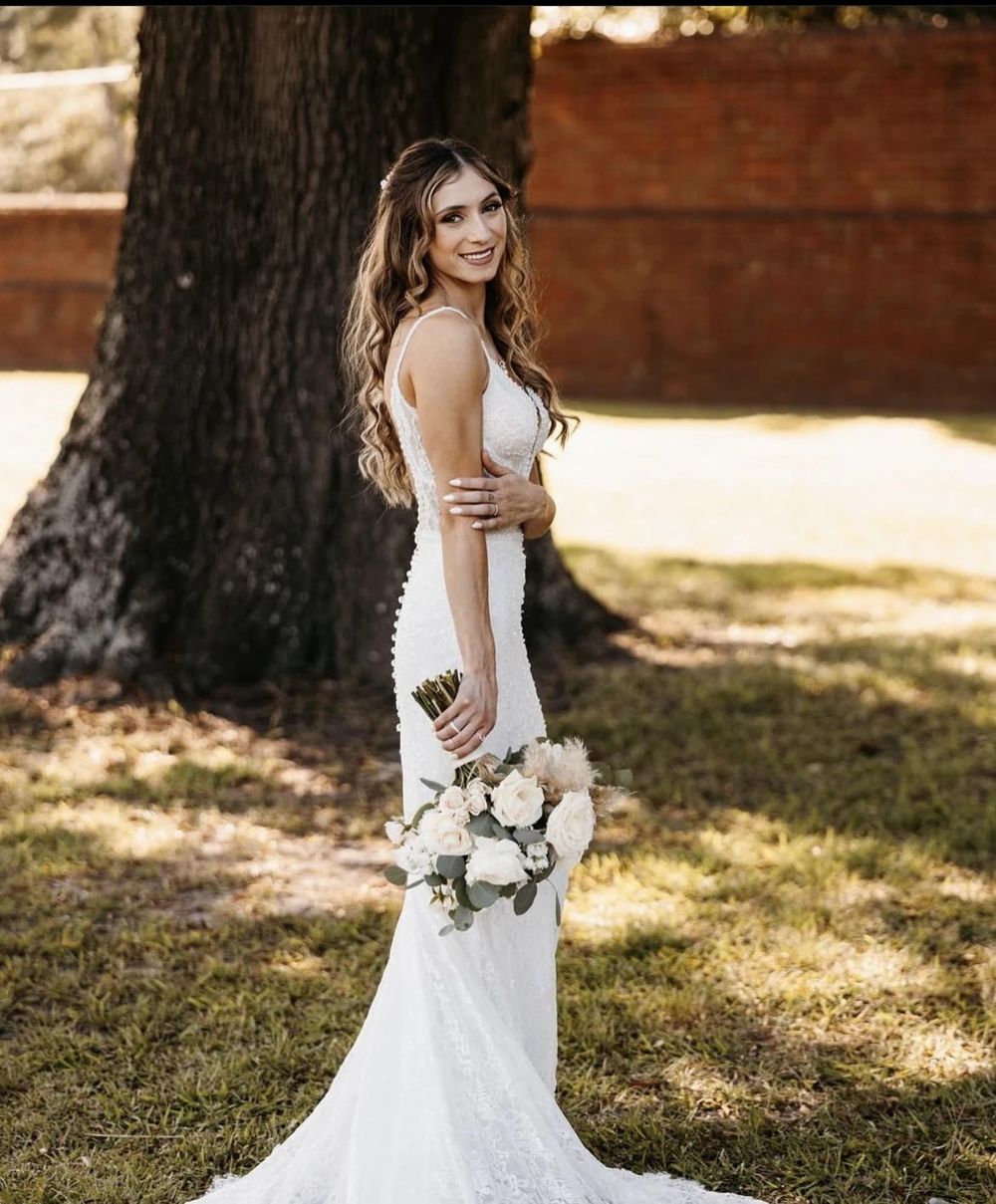 A bride in a fitted lace wedding gown poses outdoors holding a white and blush floral bouquet near a large tree.