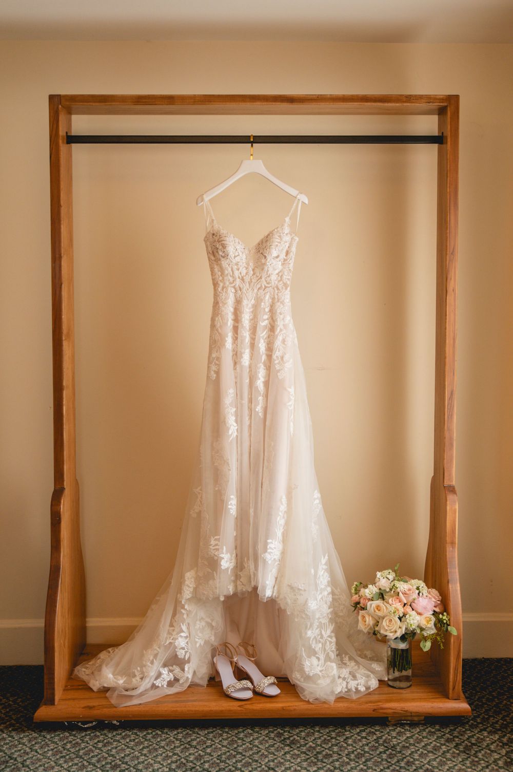 A delicate lace wedding dress hangs on a wooden frame with white shoes and blush roses below.