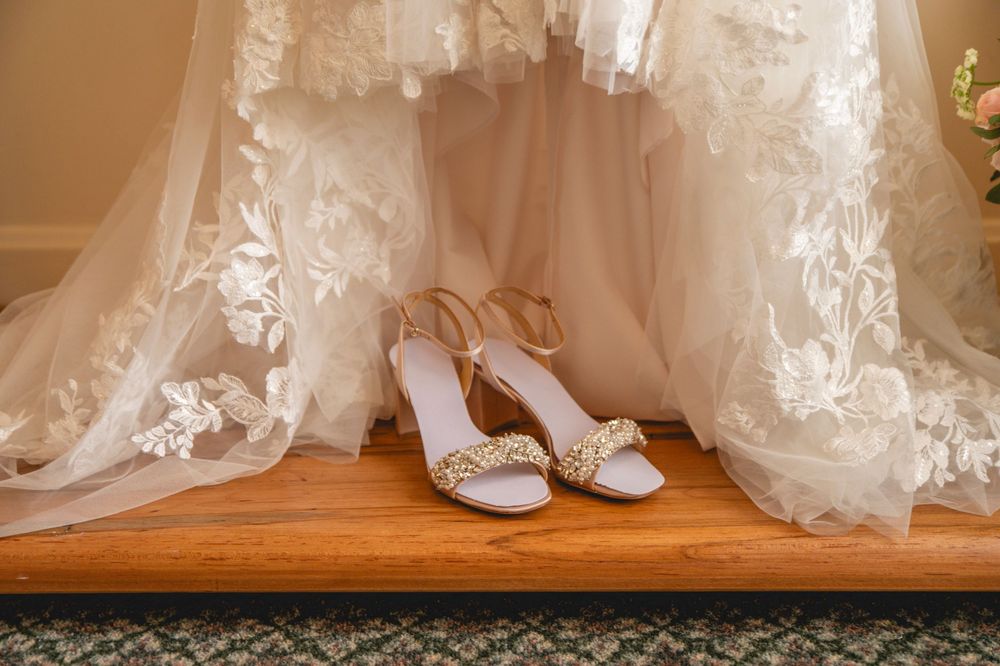 White bridal shoes with sparkly embellishments sit beneath flowing lace wedding dress on wooden surface.