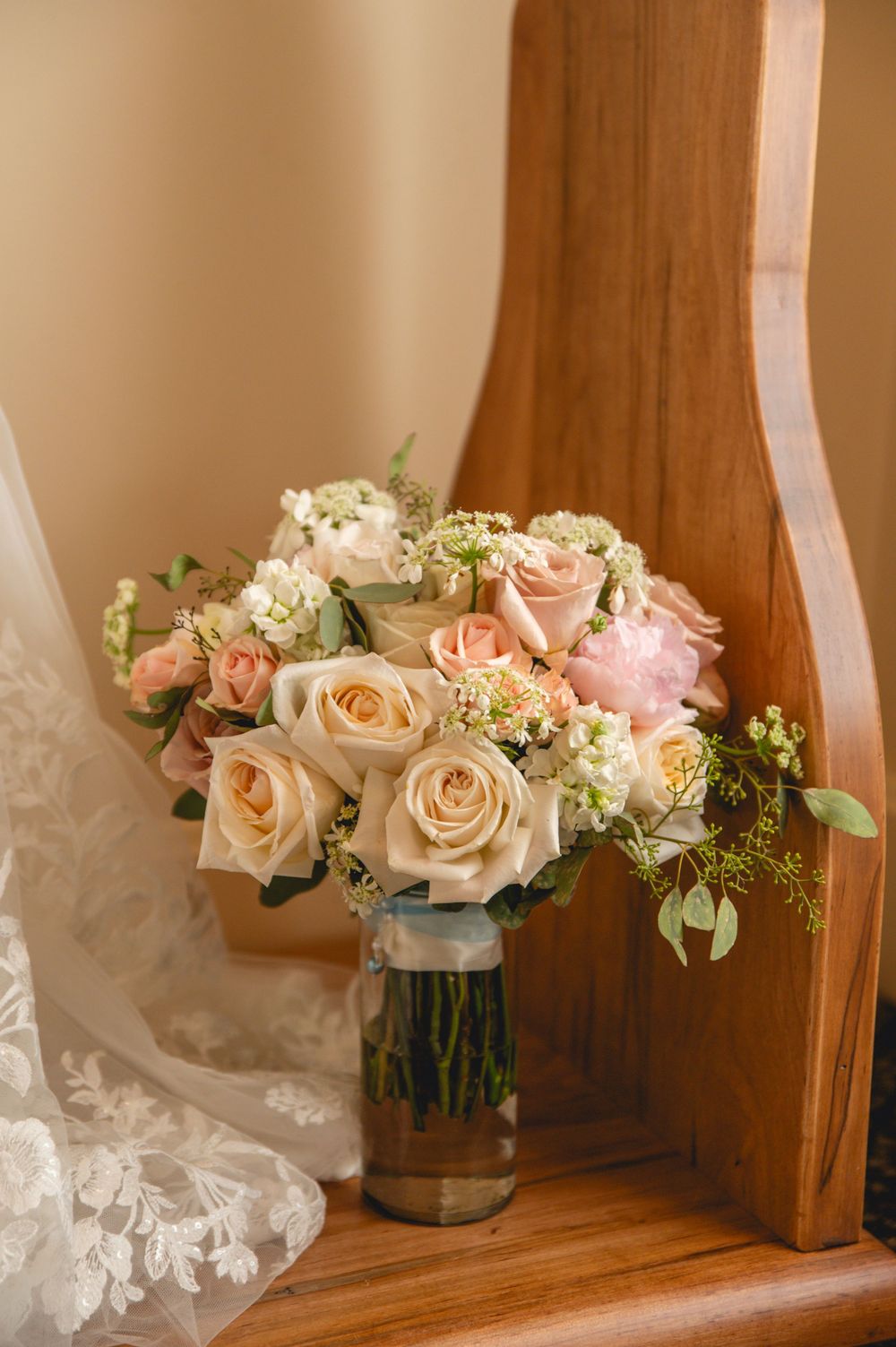 A romantic bouquet of peach and pink roses with baby's breath sits in a glass vase on a wooden surface next to lace fabric.