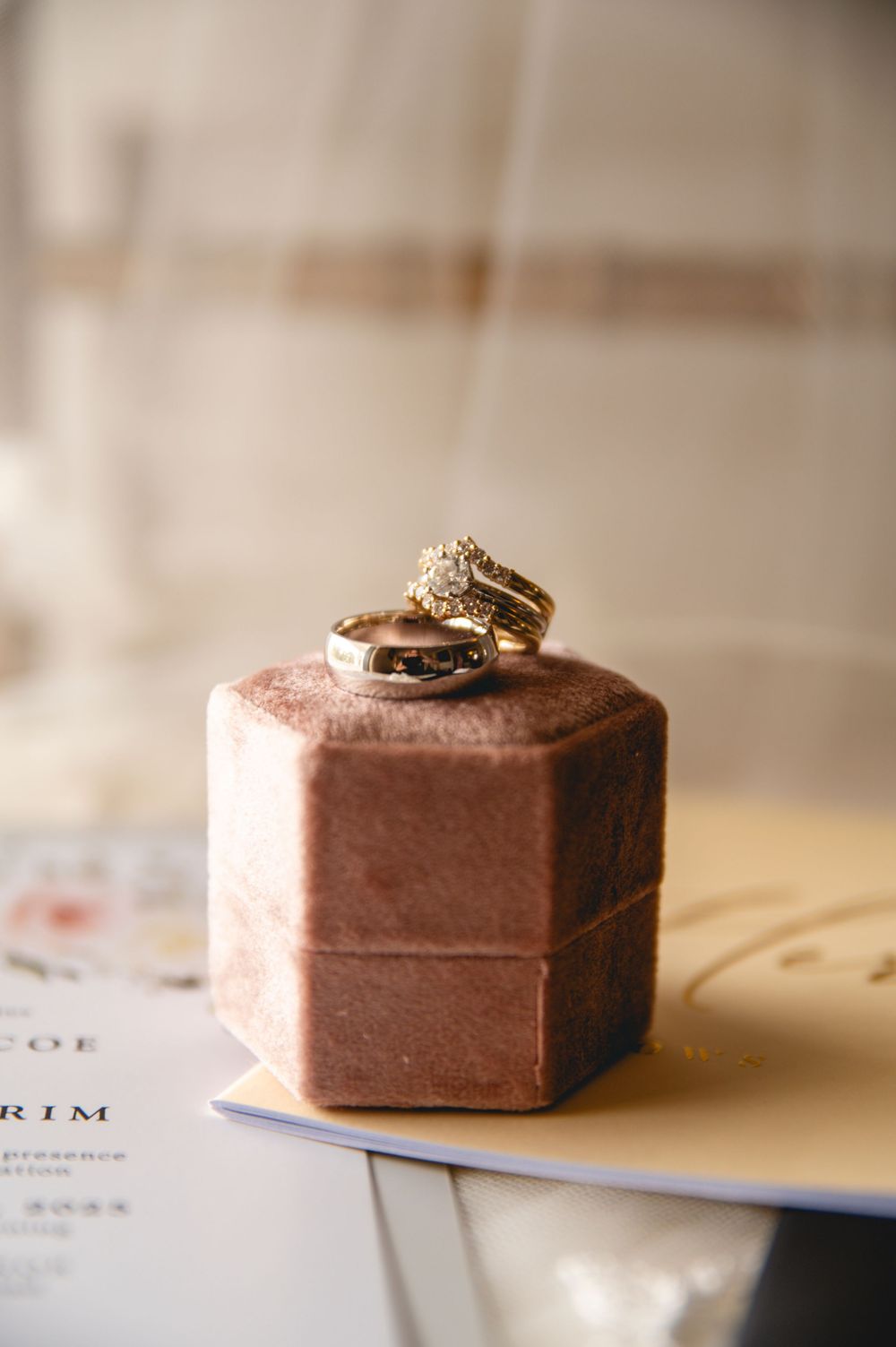 Wedding rings displayed on a pink velvet hexagonal box atop paper documents in soft lighting.