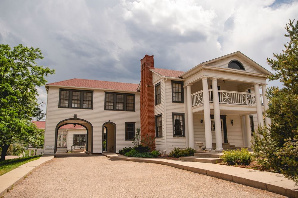 Elegant white mansion with pillared portico and curved driveway features arched carriage house and tall brick chimney.