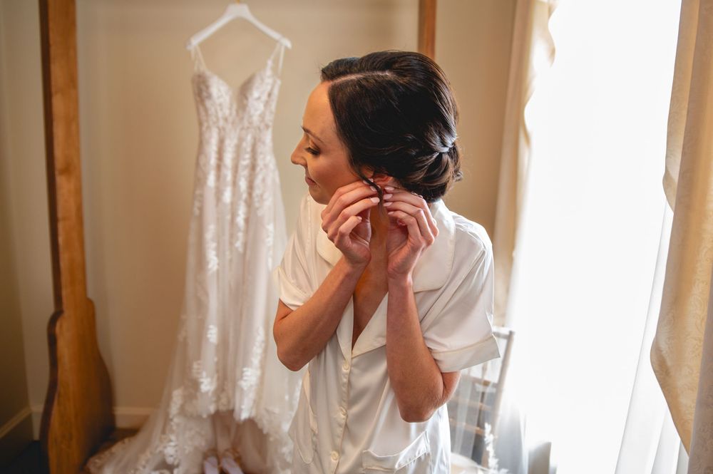 A bride looks at her white wedding dress hanging from a wooden bedpost in a sunlit room.