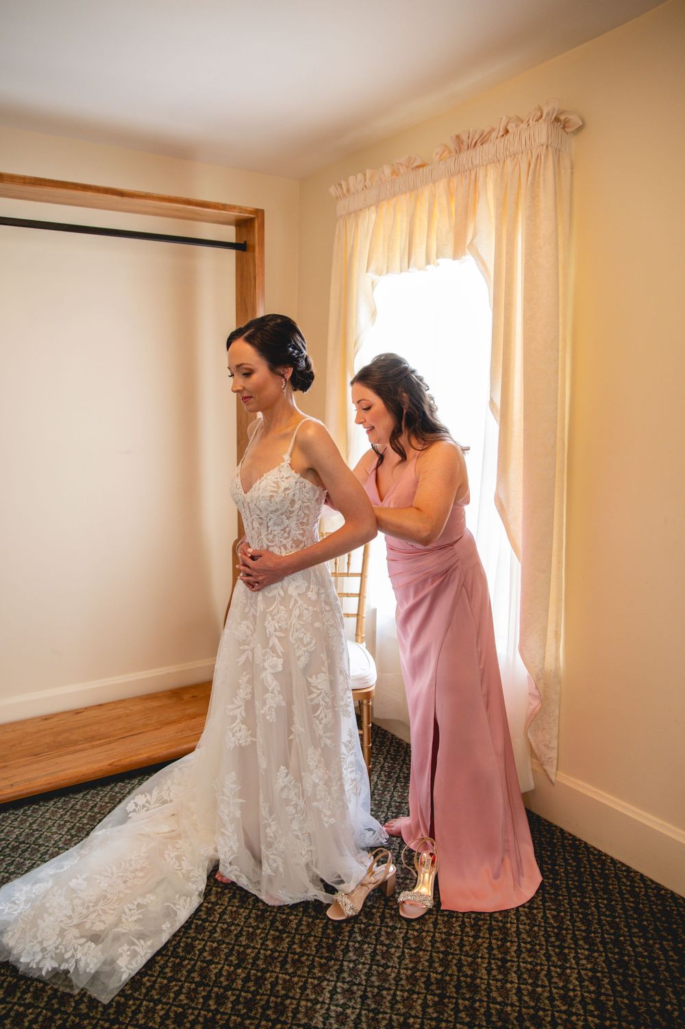 A bride in a lace wedding gown gets help with her dress from a bridesmaid wearing a dusty rose colored dress.
