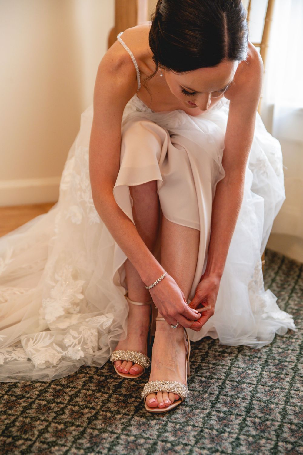 A person in a white wedding dress fastens delicate sandal straps while sitting before their wedding ceremony.