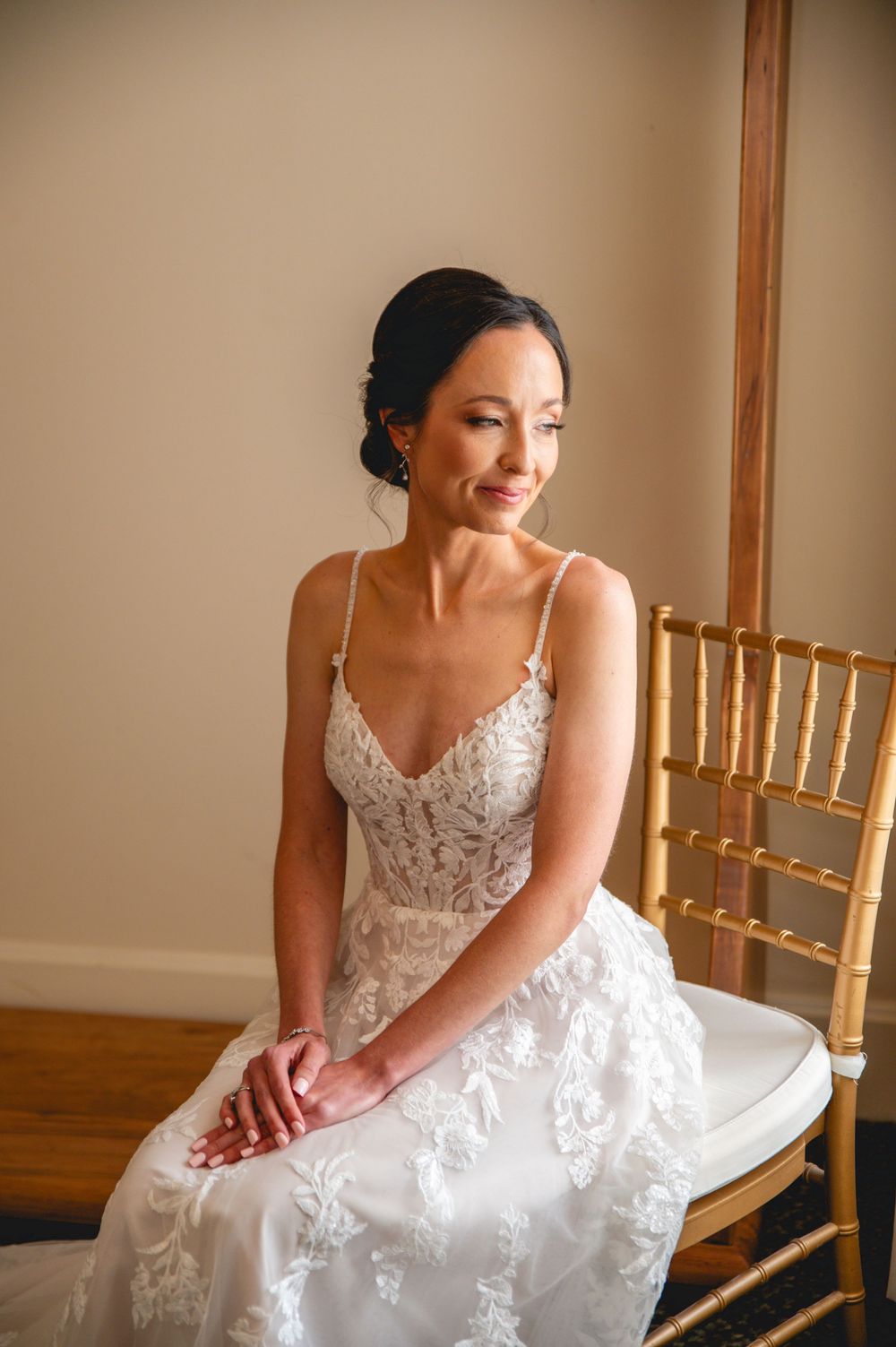Elegant bride in white lace dress sits thoughtfully in a room with gold chairs and warm lighting.