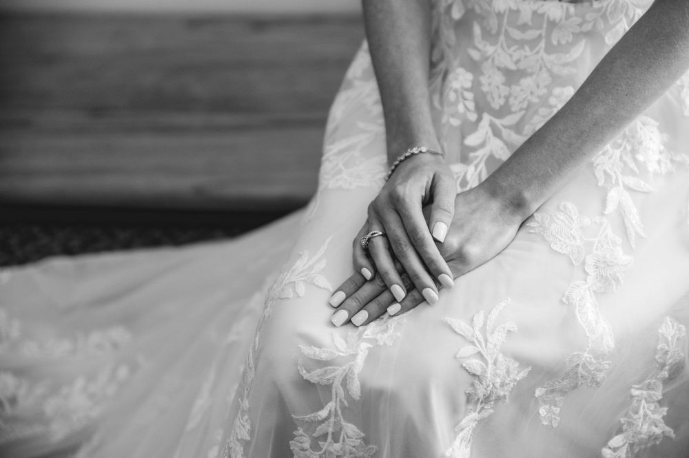 Black and white detail shot of hands resting on a white lace wedding dress with delicate floral embroidery.