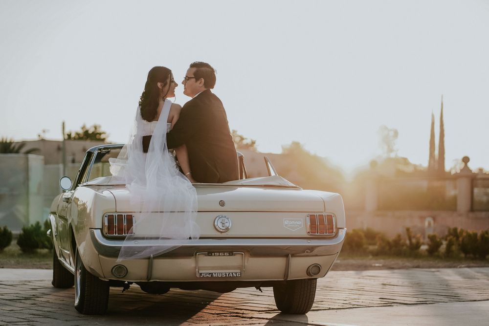 A couple sits on their classic Ford Mustang during golden hour sunset in an urban setting.