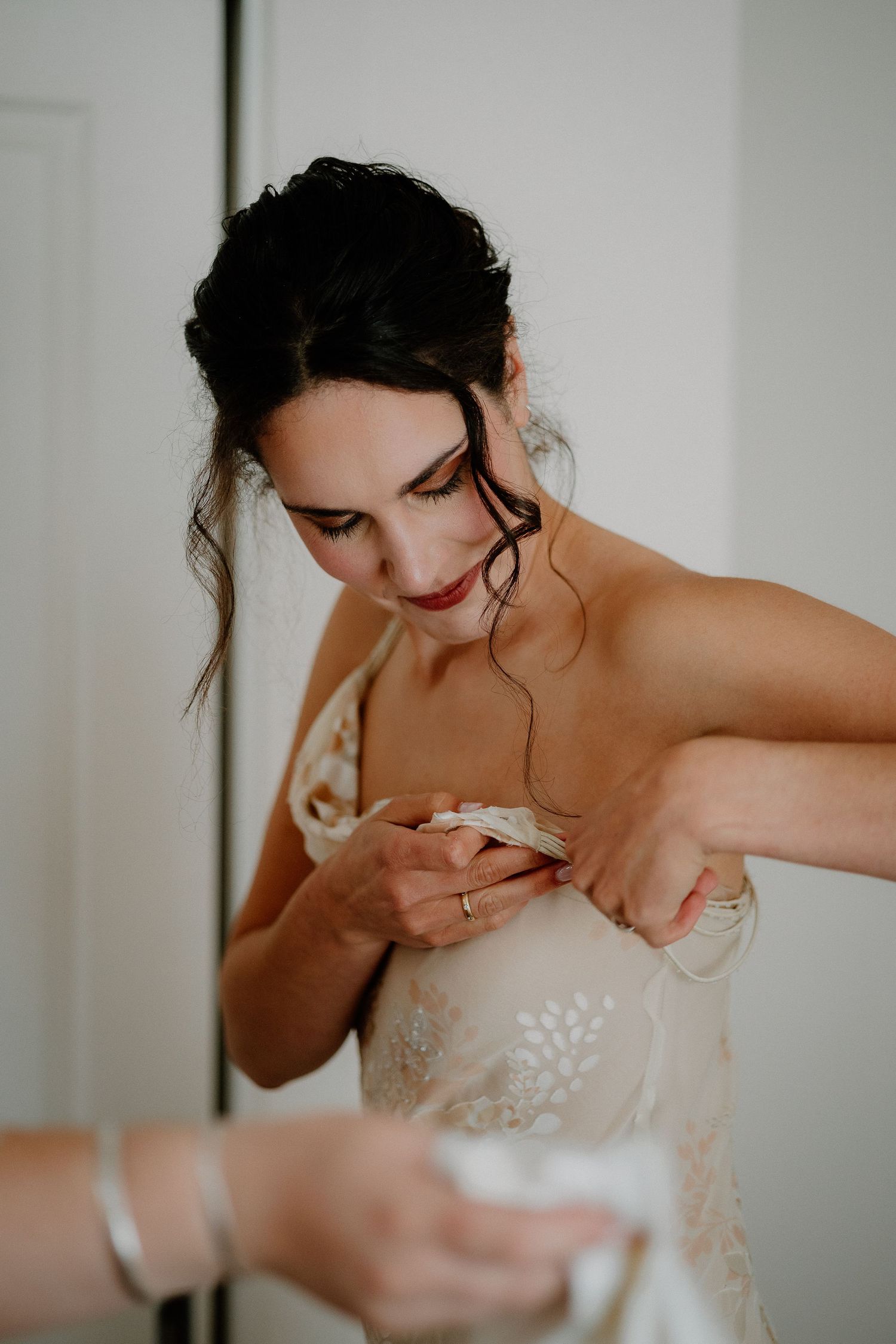 A bride in a delicate lace wedding dress getting ready, looking down at her phone with romantic loose curled hair falling.