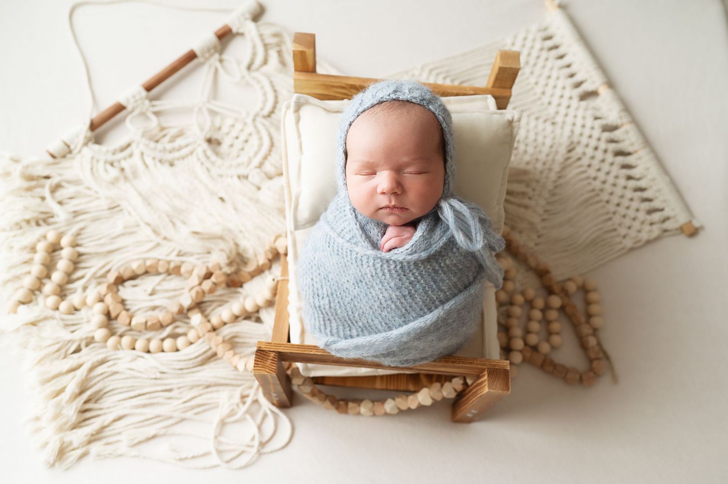 Sleeping newborn wrapped in soft gray swaddle resting in wooden basket on cream macrame backdrop.