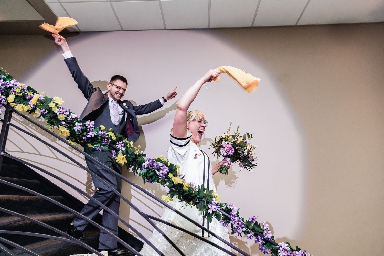 A couple celebrates joyfully with raised arms while descending a floral-decorated staircase.