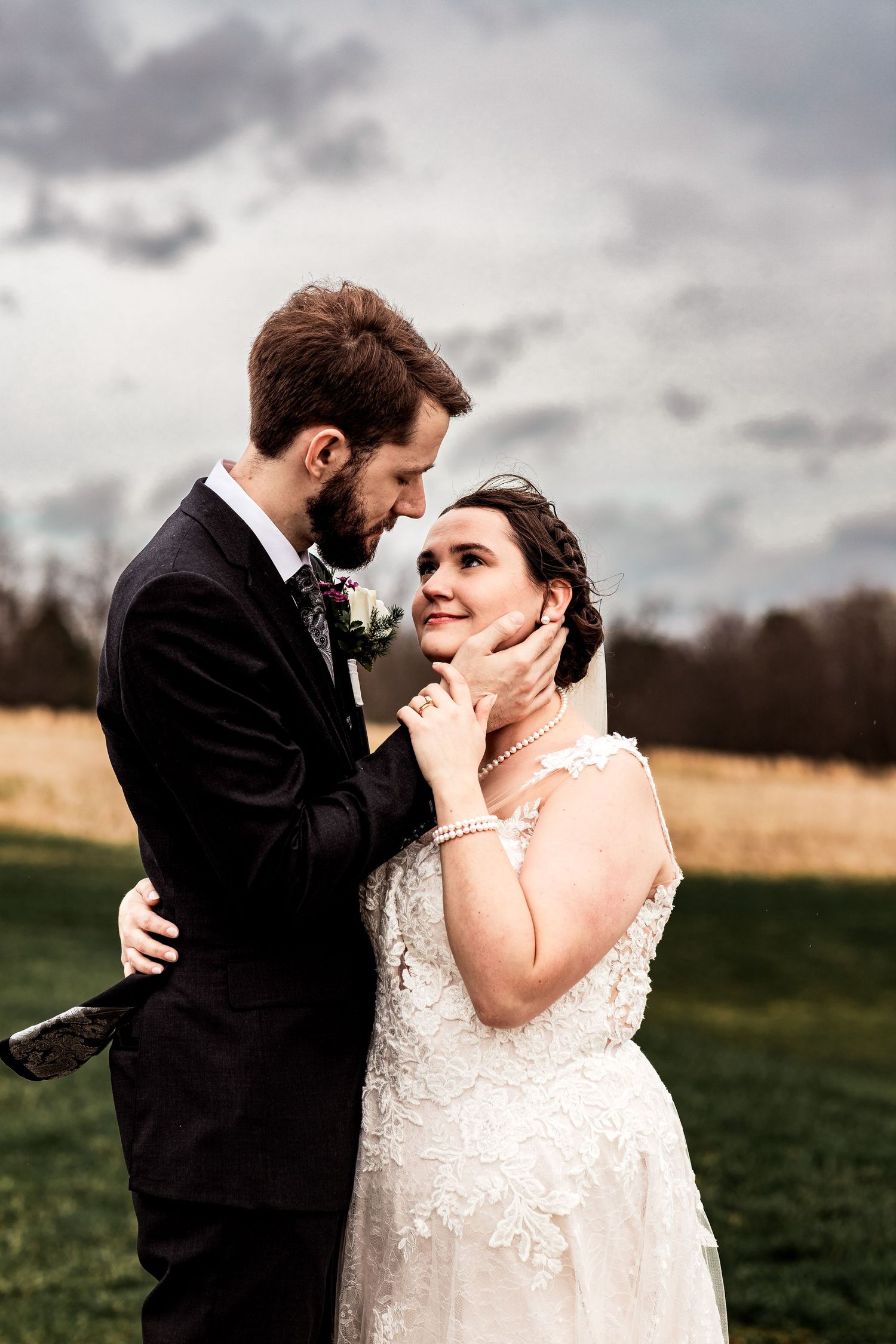 Emotional outdoor wedding portrait of bride and groom in loving embrace, standing in a field with cloudy sky background.